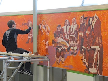 Ryan Gillis sits on a scaffold to paint in parts of a scene on the new mural at Cambridge Bay's May Hakongak Library and Cultural Centre. (PHOTO COURTESY OF THE KITIKMEOT HISTORICAL SOCIETY)