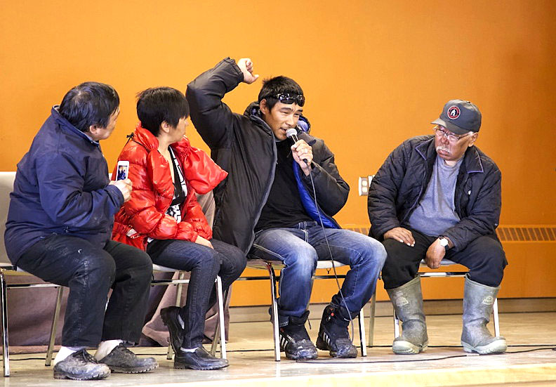 Adrian Arnauyumayuq, with microphone, demonstrates to a rapt audience in Arctic Bay how he fought off a polar bear attack with his knife early in the morning on May 22. Arnauyuymayuq and Bruce Pauloosie were sleeping in a tent overnight during a hunting trip when a bear came into the tent. It grabbed one man by the head and dragged him out while the other scrambled for his rifle. The community of Arctic Bay held a welcome home gathering at the school gym June 4 during which the two men told the audience their amazing story of survival. The bear, which was shot and killed, was skinned by the local hunters and trappers organization and the 10-and-a-half-foot pelt given to the two men. From left: Arctic Bay mayor Philip Kalluk, deputy mayor Geela Arnauyumayuq, Adrian Arnauyumayuq and HTO chair Qamajuq Oyukuluk. (PHOTO BY CLARE KINES)

