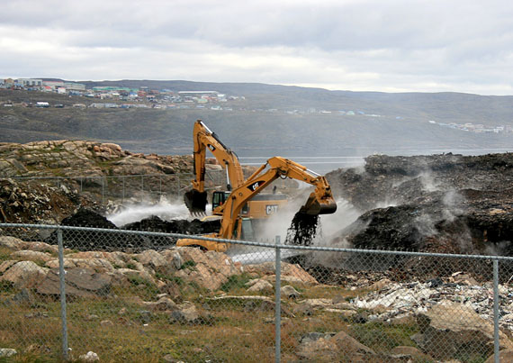 Heavy equipment operators dig into the northern end of Iqaluit’s smouldering garbage pile, Sept. 1, as unseen firefighters spray stream-fed water onto the heap. Westerly winds blow smoke from the dump fire into the city, seen in the background, and raise persistent complaints and concerns from many city residents. (PHOTO BY PETER VARGA)