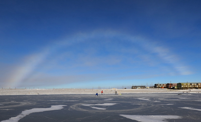 Cambridge Bay hockey players take to the ice Oct. 7 below a giant fogbow. Sometimes called a white rainbow, a fogbow forms when light hits tiny droplets of moisture in the air. (PHOTO BY RED SUN PRODUCTIONS)