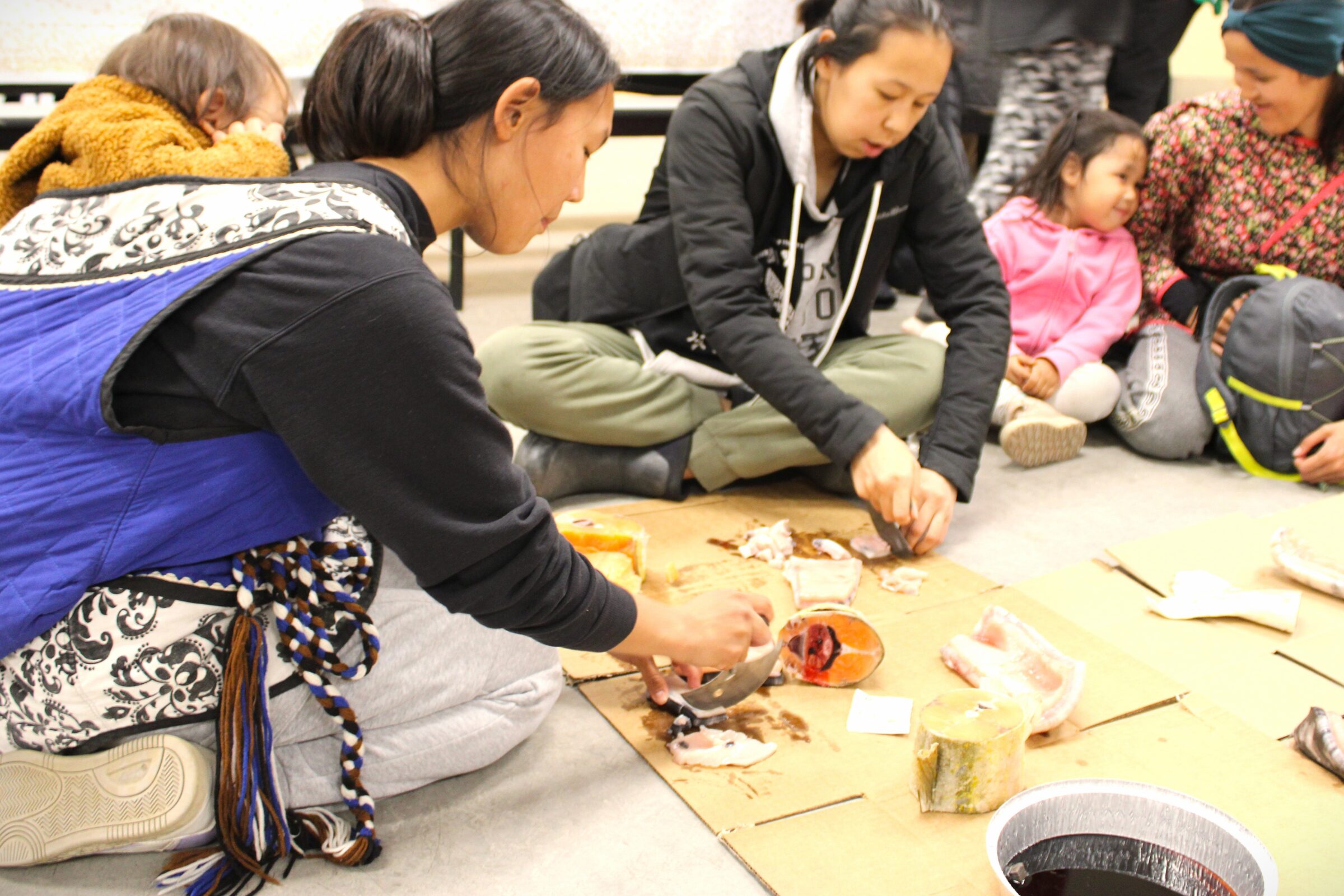 Jackson Pijamini, left, watches as his mother, Emma Pijamini, cuts beluga blubber with an ulu alongside Rose Audlakiak, Nevaeh Newkingnak and her mother Laura Newkingnak. Families enjoyed Arctic char and other country food Wednesday evening at Inuksuk High School in Iqaluit as part of the Embrace Life Council’s 20th anniversary feast. (Photo by Daron Letts)